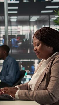 Vertical Video Focused african american general manager multitasking at desk to solve business tasks and operations on computer and documents. Woman meeting deadlines while making strategic decisions