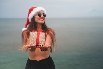 Obraz premium Banner with young brunette woman in sunglasses, red bikini and Santa hat holding gift box with bow on ocean background. Tropical Christmas and New Year in summer on island. Holiday surprise.