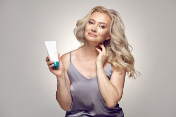 Fototapeta premium Senior woman in pajamas holding tube of cream on a grey isolated. Skin care concept.