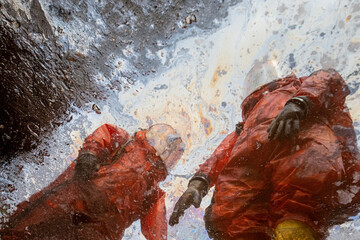 Reflection of two firefighters in hazmat chemical protective suits in an oily puddle with a rainbow sheen
