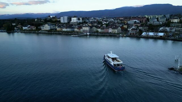 Dolly in drone shot of boat arriving in Nyon port with cityscape at sunrise in Canton of Vaud Switzerland