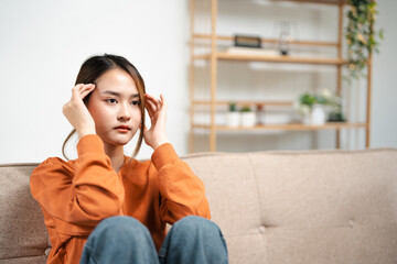 Obraz premium Upset asian woman touching forehead frustrated by problem with work or relationships, sitting on couch, embracing knees, feeling despair and anxiety, loneliness, having psychological trouble.
