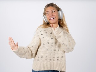 Happy Beautiful teen girl wearing white sweater sings favourite song keeps hand near mouth as if microphone wears wireless headphones, listens music