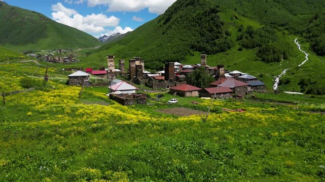 Aerial Drone Footage Captures The Ushguli Village In Georgia, Showcasing Vibrant Wildflowers On A Sunny Summer Day. The Camera Glides Over The Patara Enguri River With Majestic Mountains Behind.