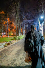 A person walking alone at night along a park pathway illuminated by streetlights. The figure, dressed in a dark coat and carrying a bag and backpack, adds a sense of mystery and solitude to the scene.