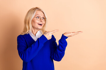 Smiling mature woman in blue jumper presenting something with a beige background, highlighting...