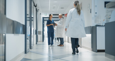 Female Nurse Briefing a Male Doctor on Patient Data in a Tablet Computer. Bright Hospital Corridor Busy with Activity as Medical Professionals in White Coats and Scrubs Walk Towards Their Departments