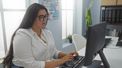 Young female working at a computer in a bright office with white walls and a notice board visible behind her