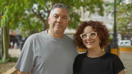 Obraz premium Middle-aged couple smiling together outdoors in a sunny urban park with lush greenery in the background.