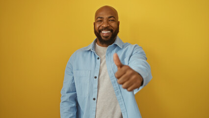 Young man smiling and giving a thumbs up over isolated yellow background wearing a light blue shirt.