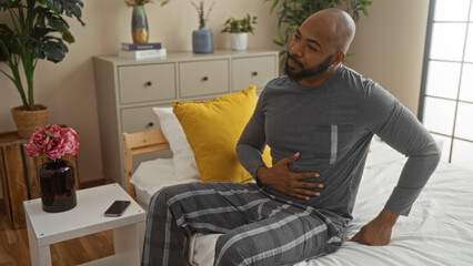 Fototapeta premium Handsome young african american man sitting on bed in cozy bedroom, holding his stomach with a concerned expression, surrounded by indoor plants and a neatly organized room