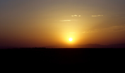 Obraz premium A stunning view of the sun setting over a dark horizon, with warm golden and orange hues blending into a serene sky. The distant silhouette of hills and the soft gradient of colors cr 