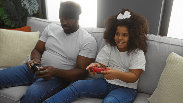 Father and daughter playing video games in a cozy living room, showcasing an african american family enjoying time together indoors, highlighting love and companionship.