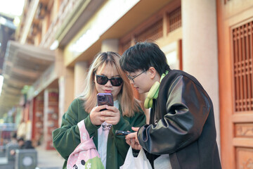 Two Chinese women in their 30s stand in front of a historical building in Jing'an District, Shanghai, China, looking at their smartphones on a cold winter day in December 2024.