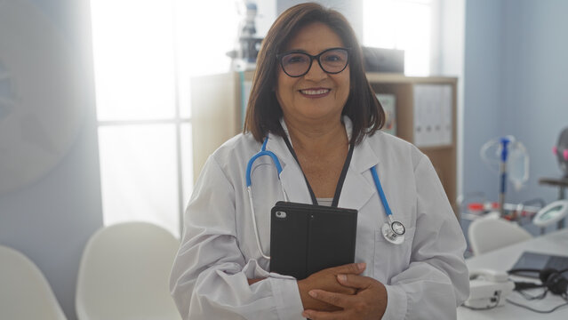 Middle-aged female doctor with short hair and glasses holding a tablet in a hospital clinic room, smiling confidently with medical equipment and white furniture in the background.