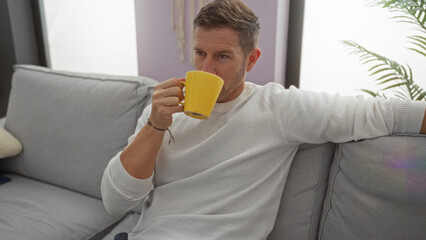 Fototapeta premium Man relaxing indoors, sitting on a couch with a coffee mug, showing a peaceful moment in a bright living room setting.