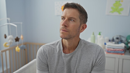 Young man in bedroom sitting near crib looking contemplative in home nursery surrounded by baby...