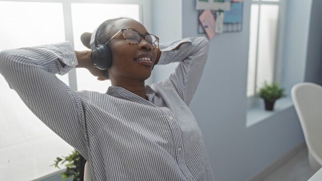 Woman relaxing with headphones in a modern office environment, looking content and comfortable, basking in the natural light from the large windows.