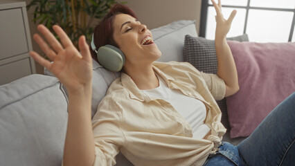 Fototapeta premium A beautiful young woman with short hair enjoying music on headphones while relaxing on a sofa in a cozy living room.