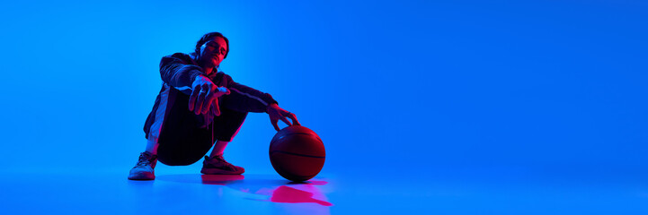 Handsome stylish young man in sport style clothes and trendy accessories posing with basketball against blue studio background in neon. Concept of sport, youth, street style, modern fashion. Banner © master1305
