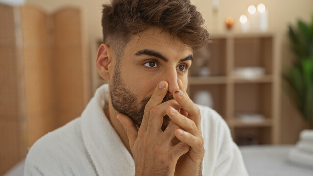 Young man with beard in spa room touching his nose and looking contemplative, surrounded by a serene wellness interior.