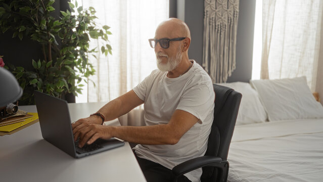 Grey-haired mature man working on a laptop in a cozy bedroom setting, creating a serene indoor working environment at home. - Powered by Adobe