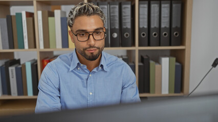 Fototapeta premium Arab man with beard and glasses working in office, surrounded by shelves with files.