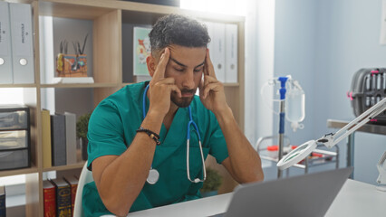 A young hispanic man in a veterinary clinic, wearing scrubs, sits at his desk with a stressed expression, highlighting the challenges of the veterinary profession.