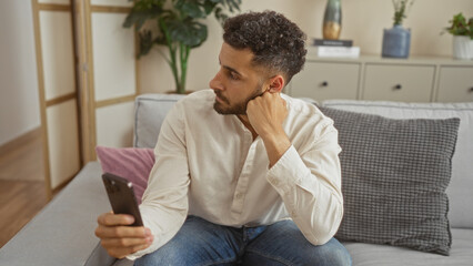 Young hispanic man sits in his cozy living room, holding a phone, with modern decor and comfortable...
