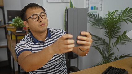 Fototapeta premium Asian man in office taking selfie with tablet, china, workplace, adult, handsome, person, attractive, indoor, man, male.