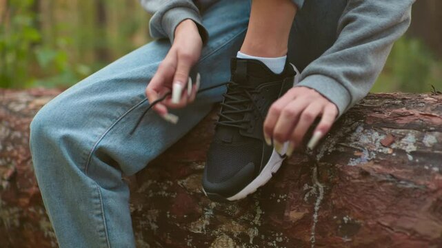 Close-up of woman with long nails tying shoelaces on black sneakers while seated on fallen tree during hike, wearing gray sweater and light blue jeans