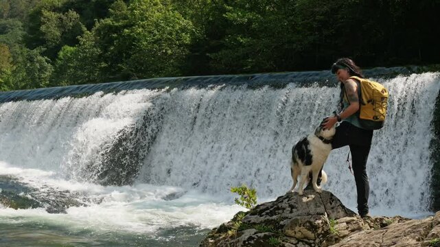 Hiker pets her border collie while standing on rocks by a waterfall. Salmon farm in the municipality of Cangas de Onis, Asturias, Spain. Tourist attraction