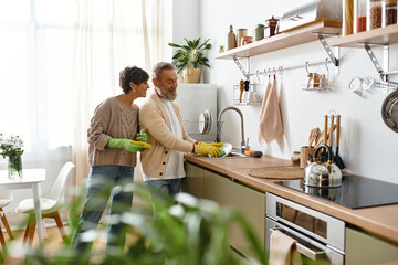 A mature couple enjoys each others company while washing dishes in a sunlit kitchen.