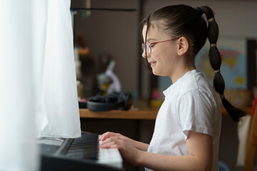 A small beautiful teenage girl in glasses with a hairstyle sitting at the piano. Blurred children's...