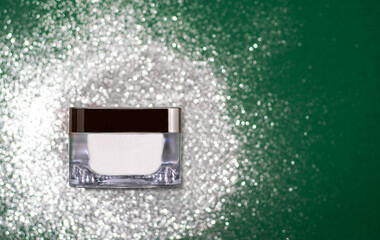Beauty face cream container on a festive green backdrop with shimmering silver glitter. For holiday skincare campaigns, luxury beauty product advertising, and Christmas-themed promotions.
