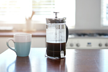 Home, kitchen and coffee plunger with cup for morning routine, drink or espresso beverage. Caffeine, mug and glass with french press on wood counter for brewing with cappuccino or latte in house.