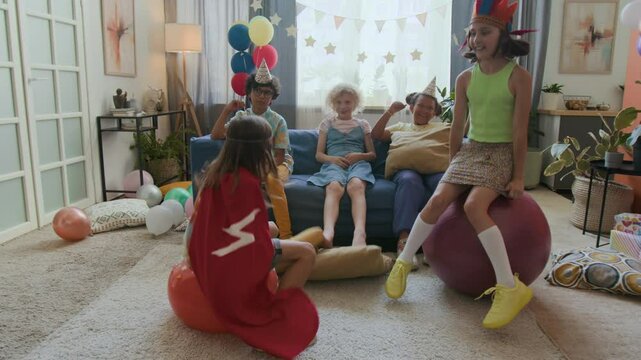 Boy wearing red cloak and girl with carnival headband bouncing on hopper balls while three friends watching them sitting on sofa in background
