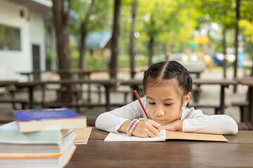 Adorable Asian girl intently writes outdoors, focused on her homework. Surrounded by books, the scene captures childhood learning, concentration, and the joy of studying in outdoor nature.