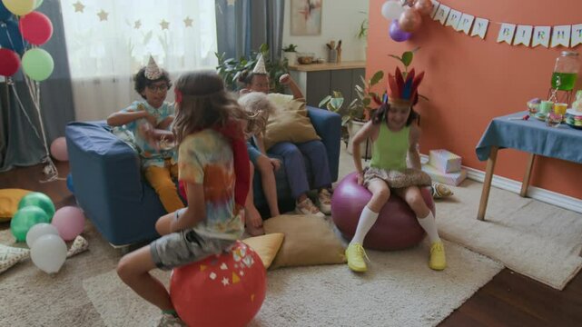 Two diverse kids jumping on bouncing balls while three friends cheering them on sofa at birthday celebration