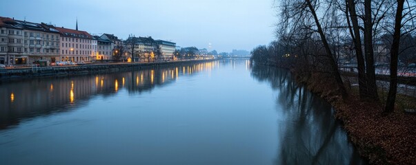 Naklejka premium A serene river scene at dusk, showcasing reflective water and illuminated buildings along the bank, framed by trees.