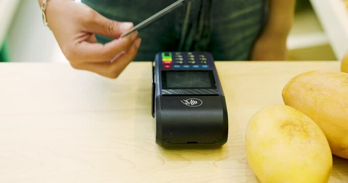 Close-up of an Asian woman tapping a credit card on a card reader for a contactless payment at a fruit shop. Fresh fruits like mangoes counter, modern retail practices. Credit card machine