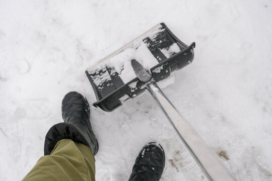 An unrecognizable person in snow boots holding a plastic shovel in order to clean, remove snow from a sidewalk for safety