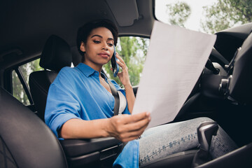 Confident young woman driving while on phone call, focusing on paperwork inside car