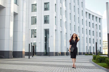 Naklejka premium A woman in a black dress walks on the sidewalk holding a laptop. She is wearing a gold necklace and has red hair. The concept of professionalism and confidence