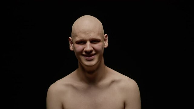 Medium closeup portrait of shirtless young Caucasian man with alopecia smiling at camera, standing on dark studio background