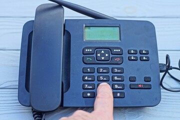 man's finger dialing a number on a modern office wireless battery black plastic push-button digital telephone with antenna and handset with charger on a wooden gray table