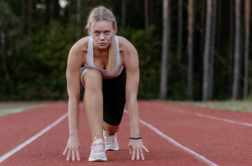 Fit, woman coach, athletic blonde with hair pulled back in a ponytail preparing for race run at start, doing sports, stretching at stadium, engaged against nature backdrop in summer outside