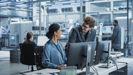 Portrait of a Young Male Assembly Line Worker and Hispanic Female Engineer Collaborating on a Project, Using Computer and Talking in a Factory with Robotic Arms Producing Modern High Tech Devices.