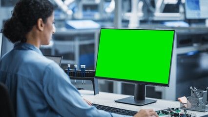 Fototapeta premium Hispanic Female Robotics Engineer Using Desktop Computer with Green Screen Chromakey Mock Up Display in Factory Office. Technological Research and Development Facility with an Autonomous Assembly Line
