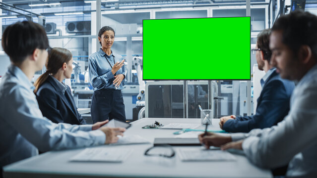Electronics Factory Office Meeting: Hispanic Female CEO Presenting Innovative High-Tech Device On TV With Green Screen Chromakey. Diverse Engineers And Technicians Listening, Planning Production.